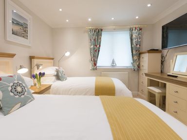 A bedroom with two beds a desk and a window with floral curtains at Surf and Sand in Carbis Bay
