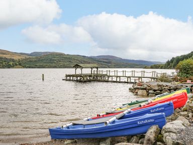 Several colorful kayaks on the shore next to a wooden dock on a lake with hills in the background at Firbush Lodge in Killin