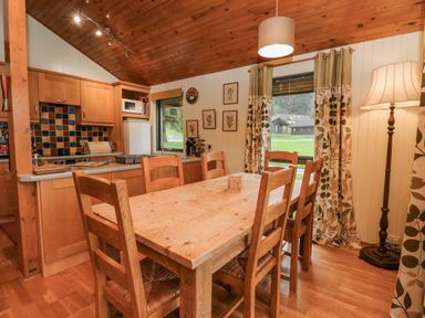 A dining area with a wooden table and six chairs next to a kitchen with wooden cabinets and two windows with curtains at Firbush Lodge in Killin