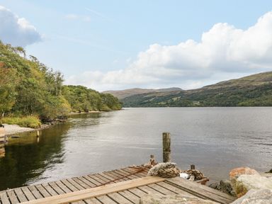 A wooden dock by a lake with trees on the shore and hills in the background at Firbush Lodge in Killin