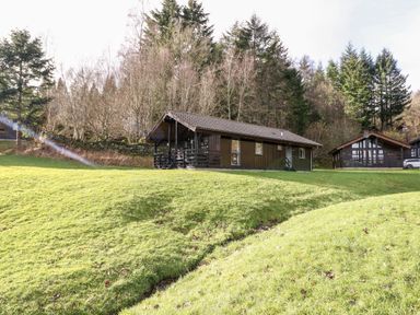 A wooden cabin on a grassy hillside with trees in the background at Firbush Lodge in Killin