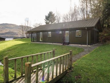 A wooden cabin with a tiled roof near a small wooden bridge over a grassy area at Firbush Lodge in Killin