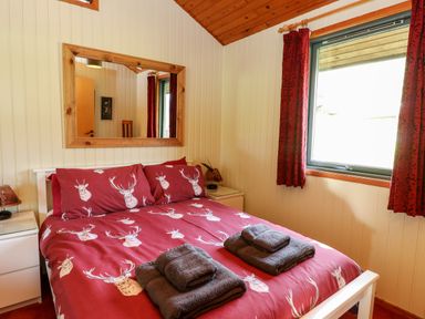 A bedroom with a double bed with deer print bedding two folded towels bedside tables and a window with red curtains at Firbush Lodge in Killin