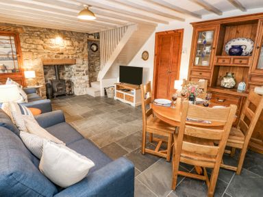 A living room with a blue sofa a stone fireplace a wood burning stove a wooden dining table and chairs and wooden cabinets at Dunnock Cottage in Broad Haven