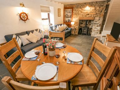 A dining area with a wooden table set for four and sofas with cushions in a living room featuring a stone fireplace at Dunnock Cottage in Broad Haven