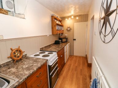 A narrow kitchen with wooden cabinets a stove a microwave and nautical decorations at Dunnock Cottage in Broad Haven