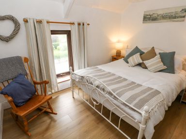 A bedroom with a bed with pillows a wooden rocking chair next to a window with curtains and a bedside table with a lamp at Dunnock Cottage in Broad Haven