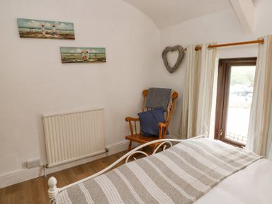 A bedroom corner with a wooden chair holding a blanket and pillow next to a window and bed at Dunnock Cottage in Broad Haven
