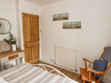 A bedroom with a wooden door a rocking chair with a cushion two paintings above a radiator and a small wooden table with flowers at Dunnock Cottage in Broad Haven