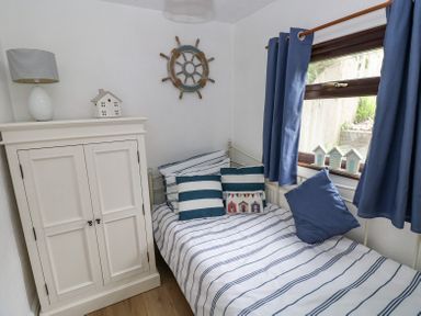 A small bedroom with a single bed with striped bedding and multiple pillows a white cabinet with a lamp and decorative house and a window with blue curtains at Dunnock Cottage in Broad Haven