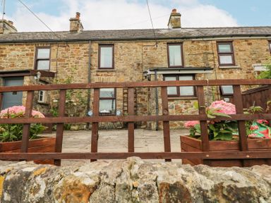 A stone house with four windows and a small patio with a table and chairs behind a wooden fence with flower pots at Dunnock Cottage in Broad Haven