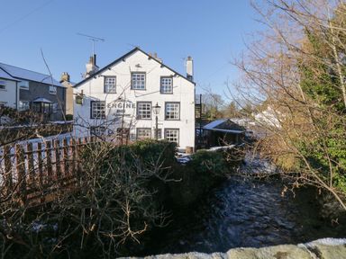 A white building with a sign for The Engine Inn Restaurant next to a river and leafless trees at Lavender Cottage in Cark In Cartmel