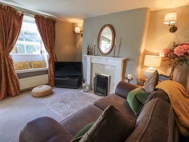 A living room with a sofa cushions and a blanket a fireplace with a mirror above a television near a window with curtains and pillows at Lavender Cottage in Cark In Cartmel