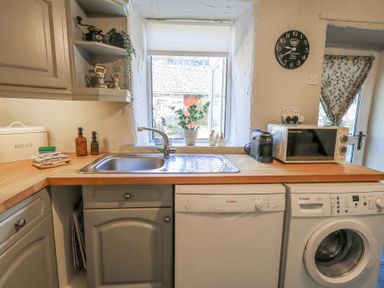 A kitchen with a sink beneath a window a bread box soap dispensers a microwave coffee machine washing machine and dishwasher at Lavender Cottage in Cark In Cartmel