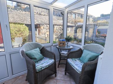 A sunroom with two wicker chairs a round table and large windows showing stone buildings outside at Lavender Cottage in Cark In Cartmel