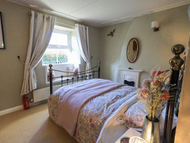 A bedroom with a bed a window with curtains and cushions and a fireplace at Lavender Cottage in Cark In Cartmel