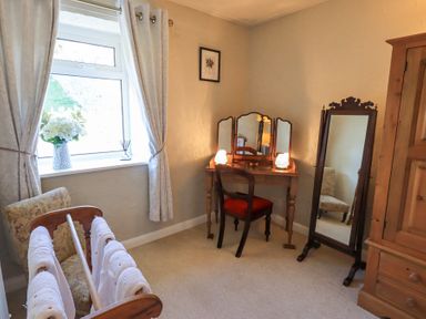 A bedroom corner with a wooden vanity table and chair a standing mirror a window with curtains and a drying rack with towels at Lavender Cottage Cark In Cartmel