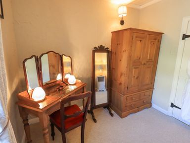 A wooden dressing table with three mirrors and two lamps next to a wooden standing mirror and wardrobe in a room at Lavender Cottage in Cark In Cartmel