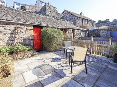 A stone patio with a round glass table and two chairs beside a stone building with a red door and a wooden fence at Lavender Cottage in Cark In Cartmel