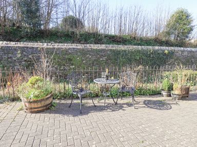An outdoor patio with two metal chairs and a round table with a bottle and glasses near plants and a stone wall at Lavender Cottage in Cark In Cartmel