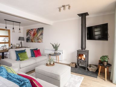 A living room with a grey sofa and cushions a wood burning stove a wall-mounted TV and a dining table at 240 Cae Du in Abersoch
