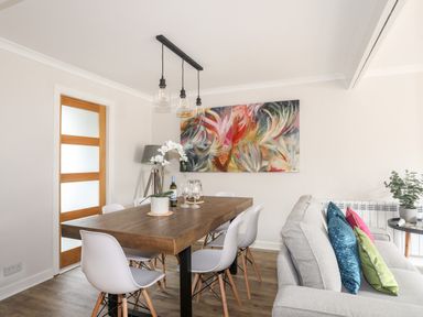 A dining room with a wooden table white chairs a gray sofa with colored cushions and abstract art on the wall at 240 Cae Du in Abersoch