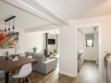 An open plan living and dining area with a wooden dining table and white chairs next to grey sofas and a wood burning stove at 240 Cae Du in Abersoch