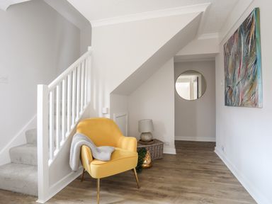 A hallway with a yellow armchair and a gray blanket near stairs a round mirror and abstract painting on the walls at 240 Cae Du in Abersoch