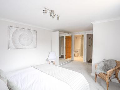 A bedroom with a bed a white chair a wicker chair with a gray throw a mirrored wardrobe and an open door to a bathroom at 240 Cae Du in Abersoch