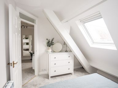 A bedroom corner with a white dresser under a slanted ceiling and a window showing an adjacent room with bunk beds at 240 Cae Du in Abersoch