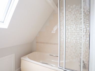 A bathroom with a bathtub under a slanted ceiling with a skylight and a glass shower screen with square patterns at 240 Cae Du in Abersoch
