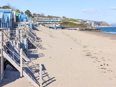 A sandy beach with beach huts on the left and buildings on a hill in the background at 240 Cae Du in Abersoch