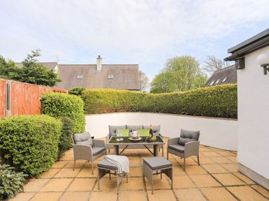 An outdoor patio with a table set with glasses and cushions on chairs surrounded by bushes at 240 Cae Du in Abersoch