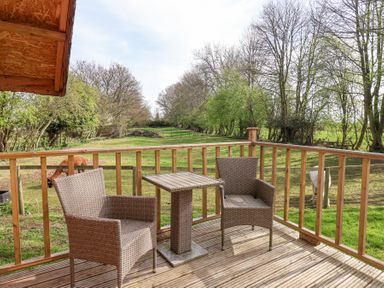 A wooden deck with two chairs and a small table overlooking a fenced grassy area with trees at Duck Lodge in Bedale