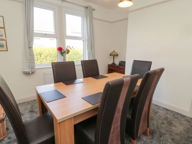 A dining room with a wooden table and six dark chairs near two windows with curtains and a small cabinet with a lamp at Bisley House in Amble