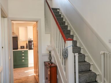 A staircase with carpet runner and a small cabinet with lamp beside it leading to a kitchen with green and white cabinets at Bisley House in Amble