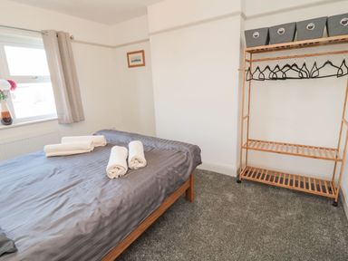 A bedroom with a bed covered in gray bedding and rolled towels on top next to a window with curtains and a wooden clothes rack with hangers and storage bins at Bisley House in Amble