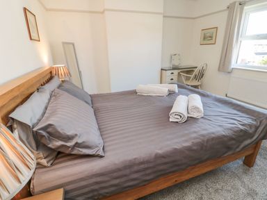 A bedroom with a double bed covered in gray striped bedding and towels on top a wooden headboard lamps a white chair and desk near the window at Bisley House in Amble