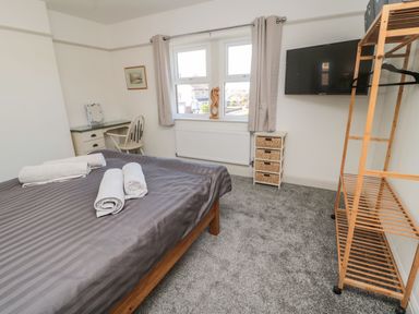 A bedroom with a bed with gray bedding and rolled towels a desk with a chair a television mounted on the wall and a wooden shelving unit at Bisley House in Amble