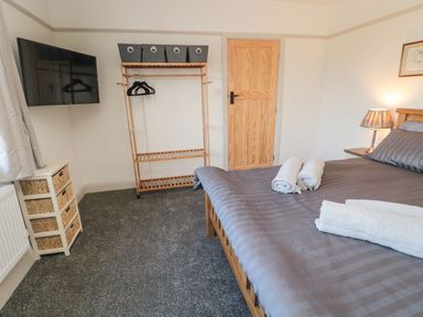 A bedroom with a bed, folded towels, a wooden door, a clothing rack with hangers and bins, a chest of drawers, and a wall-mounted TV at Bisley House in Amble