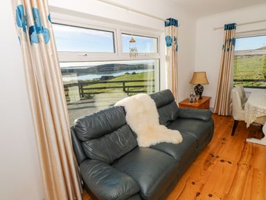 A living room with a dark leather sofa and cream curtains overlooking green hills and water at Castle Demesne House in Clifden County Galway