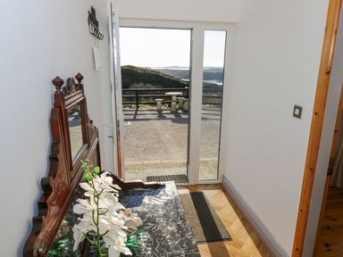 A small hallway with a wooden table with a mirror and white flowers by a glass door overlooking an outdoor seating area with hills and water at Castle Demesne House in Clifden County Galway