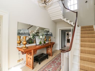 A hallway with a wooden table holding lamps and flowers next to a staircase with carpet at Rockstowes House in Uley near Dursley