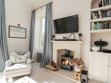 A living room with an armchair beside a fireplace with a TV mounted above and bookshelves on the side at Rockstowes House in Uley near Dursley