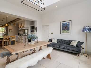 A dining area with a wooden table, bench with fur covers, a gray couch, a floor lamp, and an open kitchen at Rockstowes House in Uley near Dursley