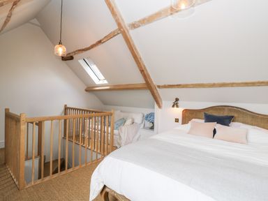 A bedroom with a large bed and pillows a small sitting area with a white armchair wooden beams on the ceiling and a stairway with wooden railing at Rockstowes House in Uley near Dursley