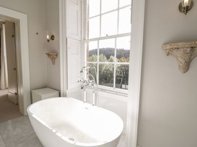 A bathroom with a white freestanding bathtub under a large window with wall sconces and decorative shelves at Rockstowes House in Uley near Dursley