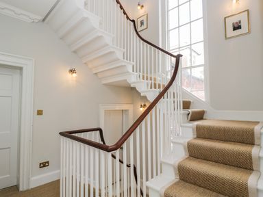 A staircase with beige carpet runner wooden handrail and white spindles next to a large window at Rockstowes House in Uley near Dursley
