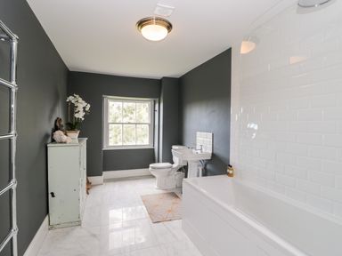 A bathroom with white bathtub white sink white toilet a window and a cabinet with flowers at Rockstowes House Uley near Dursley