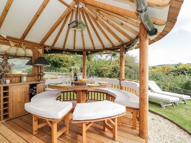 An outdoor wooden gazebo with a round dining table and cushioned semicircular bench seating at Rockstowes House Uley near Dursley